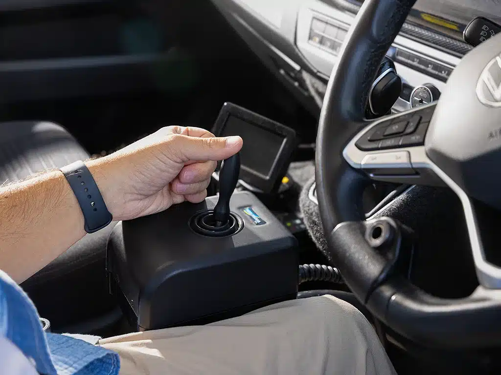 Person using an electronic joystick to control a modified driving system inside a vehicle.