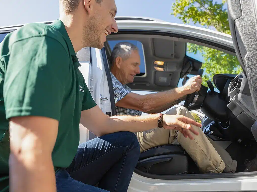 Driving occupational therapist supporting an older man during adaptive driving training session inside a modified vehicle.