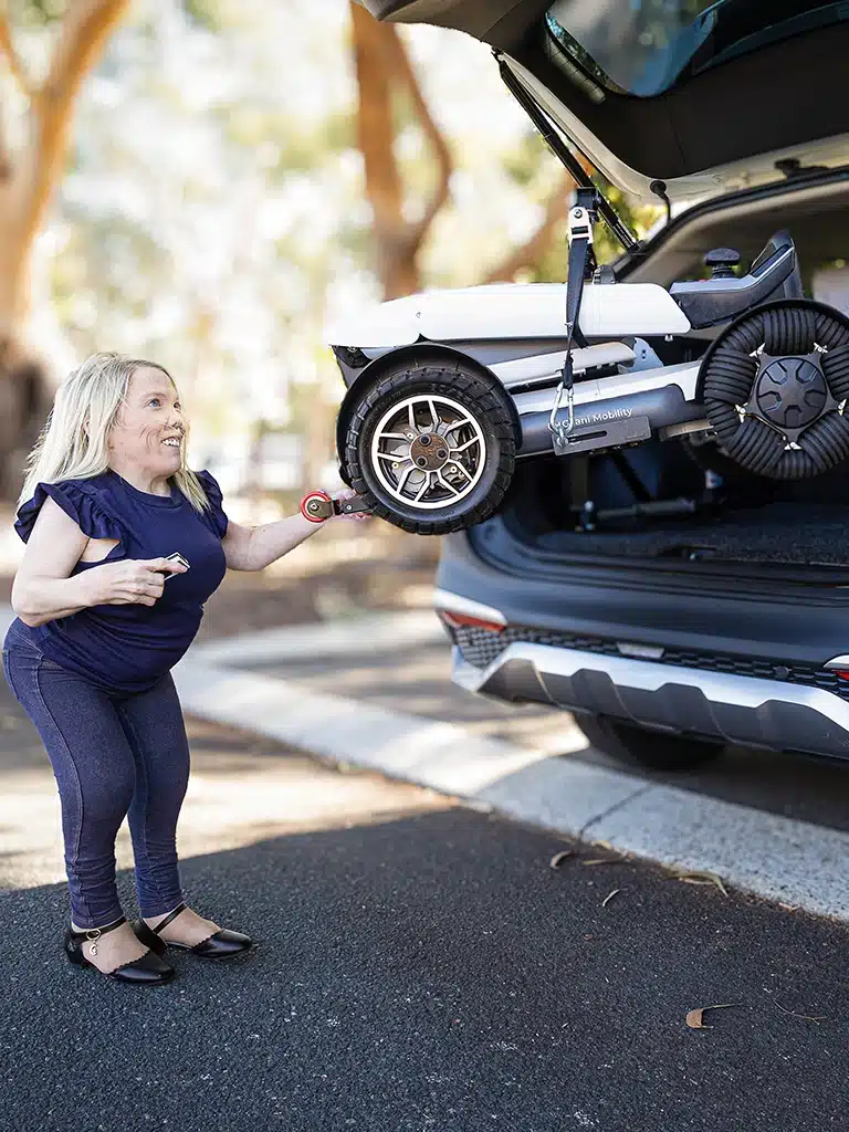Woman with short stature using an automated car boot hoist to lift a lightweight folding wheelchair.