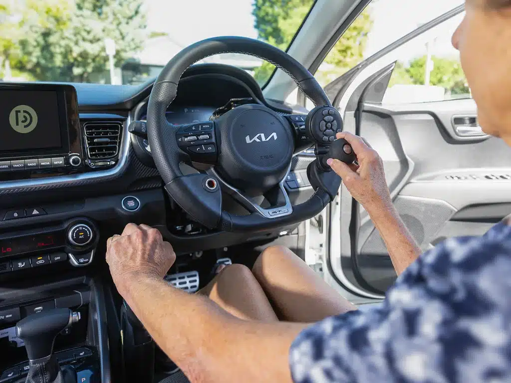Older adult using spinner knob and hand controls inside an adapted car during a driving rehabilitation session.