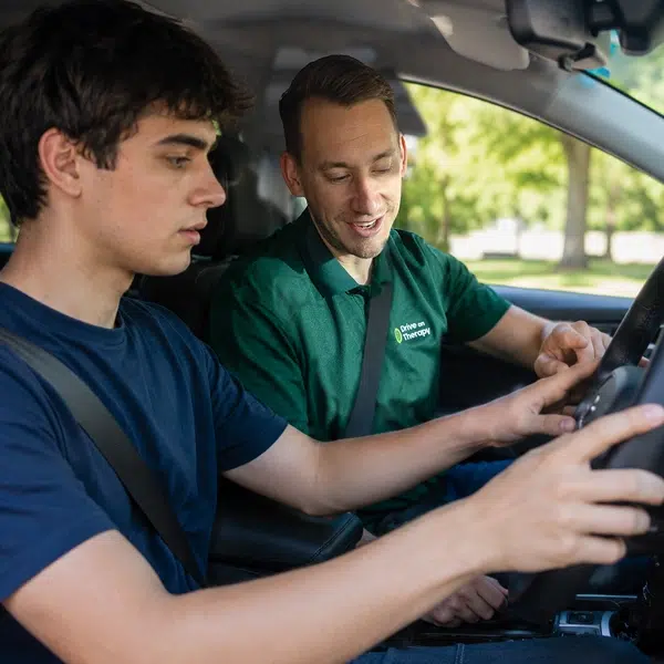 A occupational therapist providing one-on-one driving instruction to a young adult during an on-road driving assessment. Both are seated inside a dual-control assessment vehicle, with the OT offering guidance while the learner focuses on the steering wheel in a calm, supportive environment.