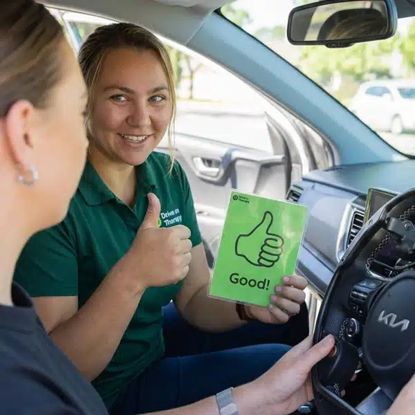 Occupational therapist gives a thumbs up while holding a green "Good!" cue card beside a learner driver.