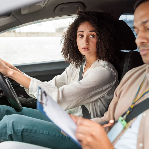 A young woman grips the steering wheel with a worried expression, glancing sideways at the driving examiner in the passenger seat who is writing on an assessment clipboard.